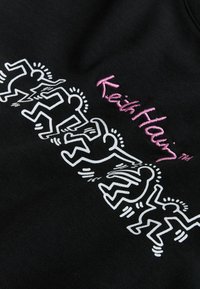 Seks hvide konturtegnede dansende menneskefigurer med strålende linjer ved siden af pink broderet tekst med teksten "Keith Haring" på sort stof.