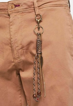 Pantaloni di cotone beige con una clip bronzo e un nappo intrecciato. Il nappo presenta perline, un pendente metallico e motivi a filo in contrasto.