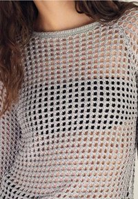 Femme portant un haut en maille crochet gris clair par-dessus un soutien-gorge noir, laissant apparaître la peau en dessous, avec des cheveux bruns ondulés tombant sur une épaule.