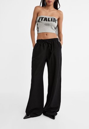 Mujer que lleva un crop top gris sin tirantes con el texto "ITALIA" y pantalones negros sueltos con cordón y zapatos negros puntiagudos.