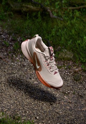 Baskets de sport beiges avec tige en mesh texturé, accents bordeaux et semelle orange. Présente des lacets et un logo swoosh sur le côté.