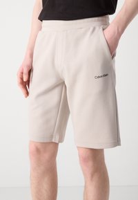 Shorts beiges en tissu doux avec une taille élastique, dotés de poches latérales et d'un petit logo noir sur la cuisse gauche.