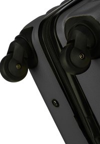 Angolo di una valigia rigida nera, con due ruote girevoli nere e una zip visibile lungo il bordo.
