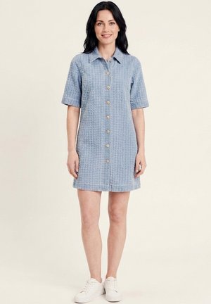 Femme aux cheveux noirs portant une robe texturée bleu clair à manches courtes boutonnée et des baskets blanches, debout devant un fond clair uni.