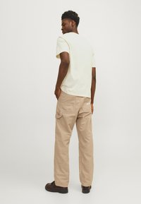 Beige cargo bukser med lommeposer, parret med en løs lys gul t-shirt, set fra ryggen. Brune sko fuldender outfittet.