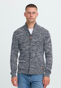 Hombre con el pelo corto vistiendo un suéter con cremallera azul marino y blanco con moteado y jeans azules, de pie frente a un fondo blanco liso.