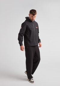 Giacca nera impermeabile con cappuccio, zip frontale e tasche. Presenta una patch rossa con logo sul petto e polsini elasticizzati.