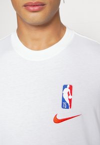 Camiseta de algodón blanca de manga corta con el logotipo de la NBA bordado en rojo y azul, y una palomita de Nike roja también bordada debajo.