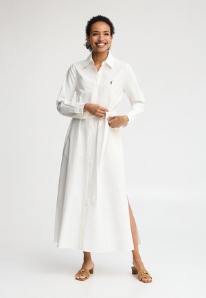 Kobieta uśmiechająca się, poprawiająca pasek przy długiej białej sukience na guziki z kołnierzykiem, rozcięciem na udzie i brązowych sandałach na obcasie, na jednolitym białym tle.