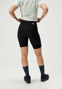 Een persoon staat met de handen op de heupen, gekleed in zwarte fietsshorts en met een patroon op de sokken, tegen een lichtgrijze achtergrond.