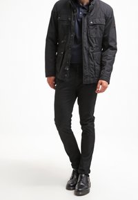 Svart, knäppt jacka med flera fickor, hög krage och matt yta, bärs över en marinblå polo och svarta byxor med stövlar.