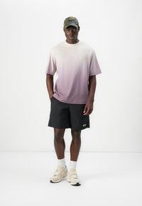 Grau meliertes T-Shirt in Helllila, kombiniert mit schwarzen Shorts und beigen Sneakers. Eine Camouflage-Kappe rundet den lässigen sportlichen Look ab.