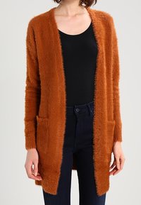 Luddig orange cardigan med öppen framsida, långa ärmar, två framfickor och ribbad struktur, bärs över en svart topp.