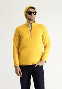 Sudadera de forro polar amarilla con cuello de media cremallera, bolsillo canguro frontal y costuras en contraste. Llevada con jeans azules y un gorro amarillo.