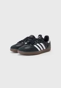 Pár černých bot Adidas Samba s bílými pruhy a hnědými gumovými podrážkami na jednoduchém světlém pozadí.