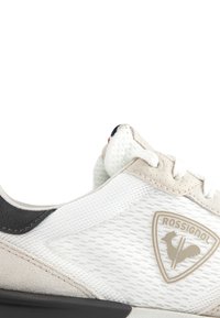 Lättviktig atletisk sko med en vit mesh-ovandel och beiga mockadetaljer, en svart hälkappa och en gummisula. Rossignol-logotyp synlig.