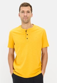 Gul kortærmet henley skjorte med rund halsudskæring, tre sorte knapper og en subtil tekstur. Har et lille logo på brystet.
