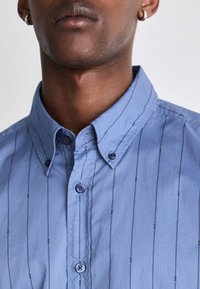 Camicia azzurro chiaro con colletto a punta, righe verticali e due bottoni sul colletto. Il tessuto sembra liscio e leggermente lucido.