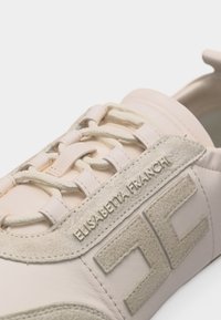 Κοντινή λήψη μπεζ αθλητικού παπουτσιού με ανάγλυφο λογότυπο και το όνομα "Elisabetta Franchi" στο πλάι, εμφανίζοντας κορδόνια και ραφές.