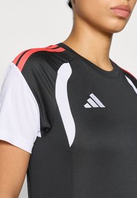 Μαύρο αθλητικό μπλουζάκι με λευκές και κόκκινες λεπτομέρειες, κοντά μανίκια και στρογγυλή λαιμόκοψη, κατασκευασμένο από αναπνεύσιμο, υφασμάτινο υλικό με υφή.