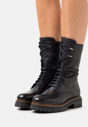 Black mid-calf lace-up boots with chunky lug soles, side zippers, and metal eyelets worn by a person with bare legs.