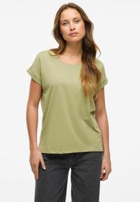 T-shirt à manches courtes de couleur vert olive en coton doux. Il présente un col rond et des manches roulées, associé à un pantalon large gris foncé.