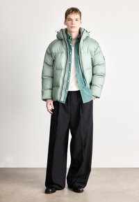 Φουσκωτό ανοιχτό πράσινο μπουφάν με κουκούλα, με φερμουάρ και τσέπες, φορεμένο πάνω από ένα πουκάμισο καρό, συνδυασμένο με μαύρο παντελόνι με φαρδιά μπατζάκια.