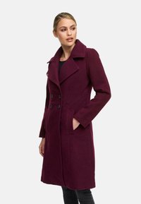 Manteau en laine bordeaux avec un design croisé, grands revers, deux poches avant et boutons noirs. Coupe ajustée et longueur genoux.