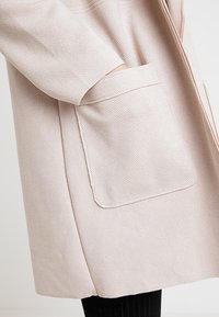 Persona che indossa un cappotto beige chiaro a trama con una grande tasca frontale e una mano parzialmente dentro la tasca.