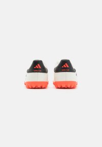 Scarpe sportive bianche con accenti neri e una suola arancione brillante. Presentano una texture liscia, un marchio evidente e tacchetti stampati per una maggiore aderenza.