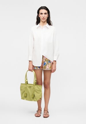 Femeie care poartă o cămașă albă cu mâneci lungi, pantaloni scurți cu model floral, sandale maro, ținând o geantă mare de mână verde deschis, stând pe un fundal simplu.