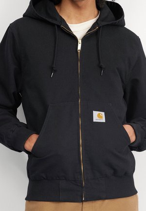 Personne portant un sweat à capuche noir zippé avec poches avant et un écusson jaune du logo Carhartt sur la poche gauche, mains dans les poches.