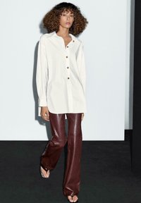 Una donna con capelli ricci indossa una lunga camicia bianca con bottoni, pantaloni in pelle bordeaux e sandali neri su uno sfondo semplice.