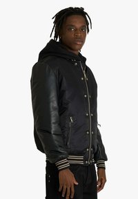 Giacca bomber nera con maniche in pelle, chiusure con cerniera e bottoni a pressione. Polsini e orlo a coste a righe, con tasche laterali, finitura opaca.