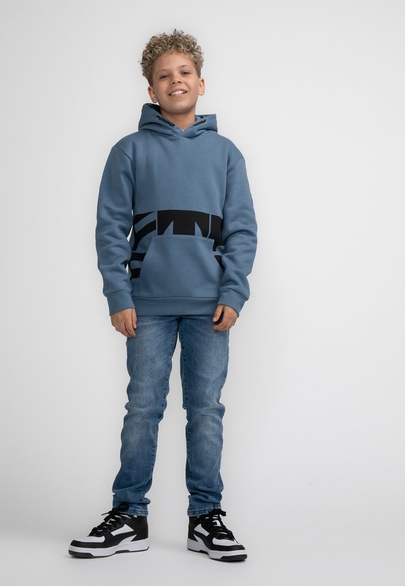 Felpa blu con una fascia grafica nera in vita, tasca frontale e cappuccio regolabile, abbinata a jeans in denim chiaro e sneakers bianche e nere.
