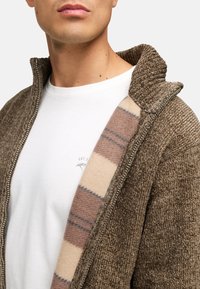 Ruskea teksturoitu takki, jossa on korkea kaulus, vetoketjukiinnitys ja sisäpuolella ruskea-beige raidallinen vuori valkoisen paidan päällä.