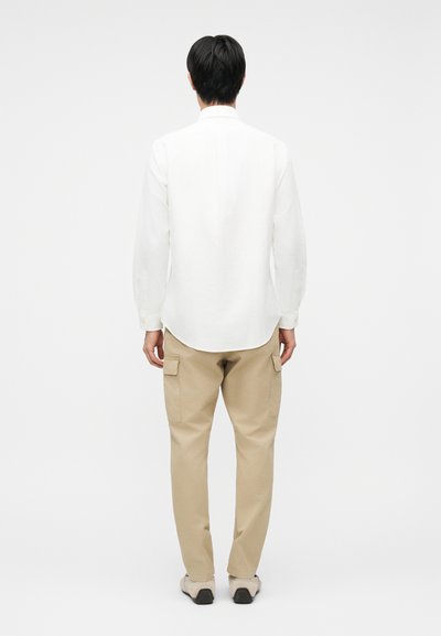 Άνδρας που στέκεται γυρισμένος με την πλάτη, φορώντας λευκό πουκάμισο με μακριά μανίκια, μπεζ παντελόνι cargo και μπεζ παπούτσια, σε απλό λευκό φόντο.