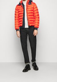 Giacca a piumino rossa con dettagli blu navy e polsini a coste, abbinata a jeans scuri e scarpe nere. Texture liscia, chiusura frontale con zip e tasche.