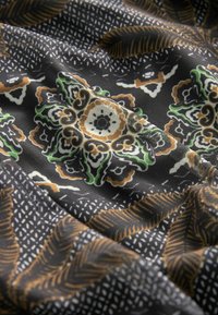 Tissu noir présentant des motifs floraux et de feuilles complexes en vert, marron et blanc. Surface texturée avec des formes et des détails variés.