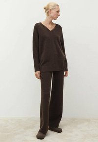 Brauner Strickpullover mit V-Ausschnitt, kombiniert mit weit geschnittenen braunen Hosen. Weiche Textur, lockere Passform und passende braune Hausschuhe.