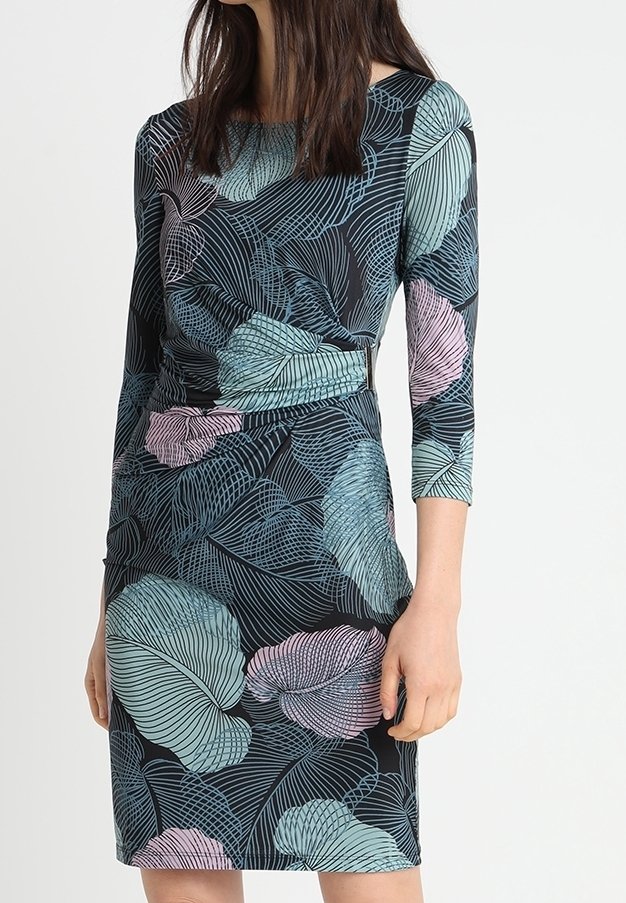 Femme portant une robe ajustée à manches trois-quarts avec un motif de grandes feuilles bleues et violettes sur un fond sombre.