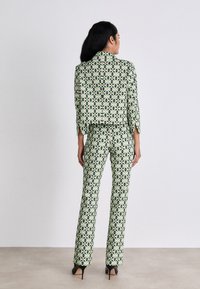 Gemusterter Anzug in Hellgrün mit geometrischen Formen, bestehend aus einer kurzen Jacke und enganliegenden Hosen. Akzentuiert mit einer glänzenden Textur und spitzen Absätzen.