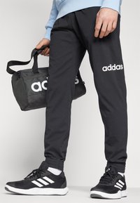 Svart joggebukse med hvitt "adidas"-logo, sammen med svarte treningessko med hvite striper. En svart treningsbag blir holdt i hånden.