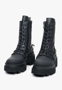 Zwarte leren combatboots met dikke rubberen zolen, vetersluiting aan de voorkant en ritssluiting aan de zijkant. Gladde textuur met matte afwerking.