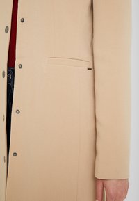 Abrigo beige de tela texturizada con acabado suave, con botones de presión y un pequeño bolsillo lateral en el lado izquierdo.