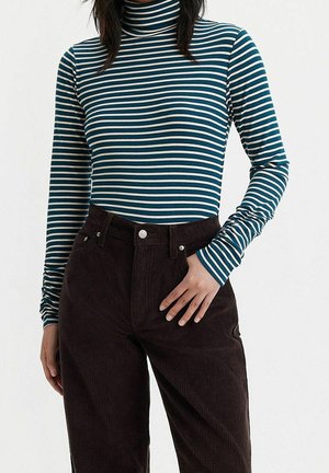 Personne portant un col roulé rayé turquoise et blanc et un pantalon en velours côtelé marron foncé taille haute, debout avec une main posée sur la hanche.