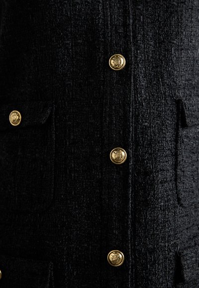 Giacca nera con una texture caratteristica, dotata di bottoni a forma di testa di leone dorati e due tasche frontali. Il tessuto ha un aspetto intrecciato, che aggiunge profondità al design.