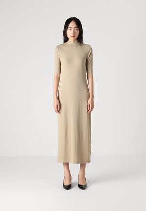Beige ribbet midi kjole med høy hals og korte ermer, med sidelommer. Modellen har på seg svarte lakkledde hæler.