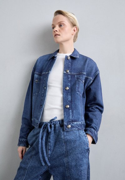 Jachetă din denim de culoare albastru închis, cu nasturi argintii și două buzunare frontale, asortată cu pantaloni din bumbac deschis, cu șnur. Țesătura pare moale și texturată.