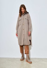 Manteau à carreaux boutonné dans des tons beiges et bruns, doté de deux poches poitrine, d'une capuche à cordon de serrage et d fentes sur les côtés ; associé à un pantalon en simili cuir marron et des bottes.